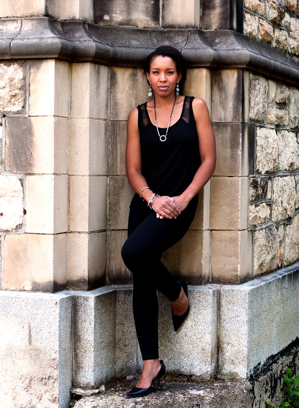 The Live Fiercely Coach | Consultant | Speaker stands leaning against a stone wall with a fierce yet welcoming look on her face. One of her legs is bent with her foot resting on the wall. Her hands are clasped resting on the bent leg.